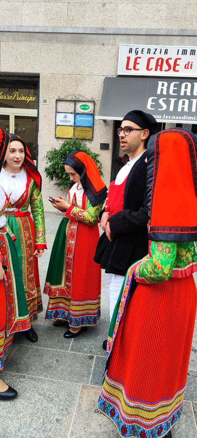 The image shows a group of people standing on a stone-paved street in the city of Sanremo, Italy. Three women are wearing traditional Sardinian clothing, including red and green skirts, embroidered blouses, and red headscarves with black lace trim. One of the women is looking at her mobile phone. A man stands next to the women. He wears a black beret, glasses, and a traditional Sardinian jacket and vest. The building in the background has a sign for "Agenzia Immobiliare Le Case di". Another sign on the building is for "Parolini" and "Sanremissimo." The street address "33" is visible on the building. "Carro Principe" is written on the storefront on the left.