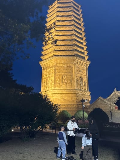 The image shows a tall pagoda-style building illuminated against a dark blue sky in Jinzhou, China. The building has multiple tiers and is detailed with carvings. Several people are standing in front of the structure, seemingly taking photos. There are trees and bushes surrounding the pagoda. The ground appears to be a mix of grass and paved surfaces. The photograph was likely taken at night due to the darkness of the sky and the illumination of the building.