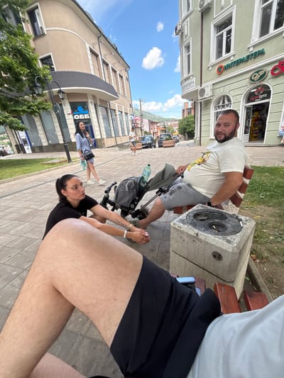 In Karlovo, Bulgaria, on a sunny day, a man and a woman are seen on a sidewalk. The man is sitting on a bench to the right, wearing a light-colored t-shirt and gray shorts, with a baby stroller beside him. He is looking towards the camera. The woman is sitting on the ground in front of the bench, wearing a black t-shirt and shorts, with glasses, and she is looking to her left. The background shows buildings with storefronts and a tree. A stroller is positioned between the man and the woman. A stone ashtray with cigarette butts is on a pedestal in front of the bench. The scene is captured on a paved walkway with buildings and a street in the distance.