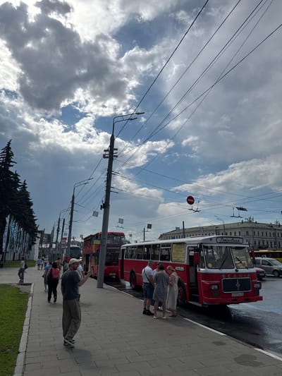 A group of people are standing on a sidewalk next to a red and white bus in Заволжье, Russia. The bus has the word "РетроВояж" written on the front. The sky is cloudy.