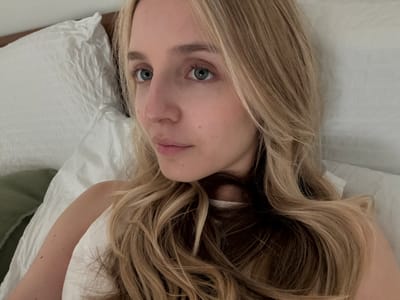 A close-up, overhead, selfie-style shot shows a young Caucasian woman with blonde hair lying in bed. She is on her side, facing left, with her head resting on a white pillow. Her eyes are open and looking to the left. She is wearing a white sleeveless top. Her hair is long and wavy, falling across her shoulders and chest. The background consists of white bedding and a corner of a green pillow on the left. The image appears to have been taken indoors, likely in a bedroom. The lighting is soft and natural, suggesting it may be daytime. There are no visible landmarks or features to indicate a specific location in Miami, United States of America.