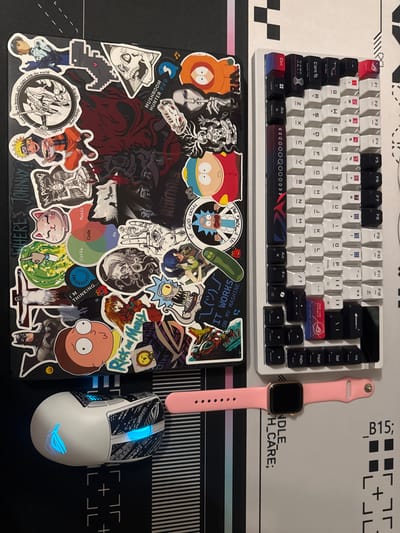 This is an overhead shot of a gaming setup on a desk. The scene is not in Kremenchuk, Ukraine. In the center of the frame is a black laptop heavily decorated with various anime and pop culture stickers. To the right of the laptop is a white mechanical keyboard with a mix of black and white keycaps, some of which have red accents. Below the laptop is a white gaming mouse with blue LED lighting. To the right of the mouse and below the keyboard, a pink smartwatch is laid out on the desk. The desk surface appears to be a black mousepad with white graphic elements.