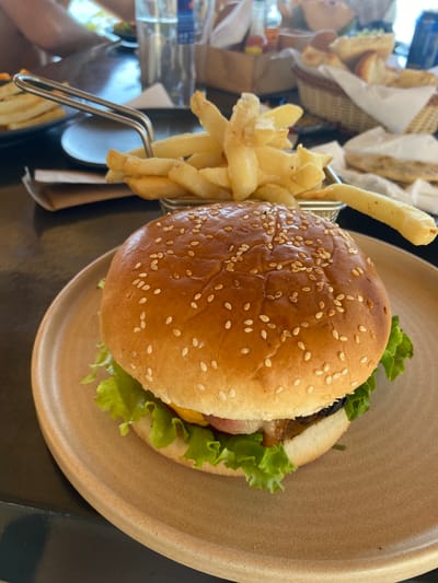 An angled top-down shot reveals a sesame seed bun hamburger with lettuce on a tan plate. A metal basket brimming with french fries sits adjacent to the burger. Other baskets filled with similar items, possibly sandwiches, are visible in the background. A bottle of water is to the left, and the scene is set on a dark table.