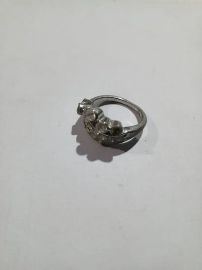 A silver-colored ring, embellished with multiple clear, faceted stones, rests on a plain white, untextured surface. The ring's design features a prominent cluster of five larger, round-cut stones arranged in a slightly curved row, flanked by smaller clear stones set along the band. The metal appears somewhat worn and dulled. A distinct shadow is cast by the ring to its bottom-right, indicating illumination from the upper-left. No other objects or individuals are present in the frame. The scene is located in the city of Phakeo, Laos.