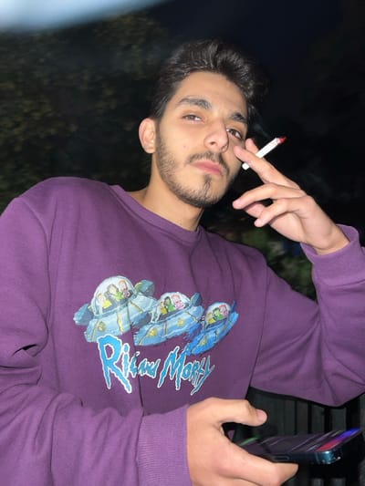 The image features a young man, likely in his late teens or early twenties, photographed at night in Al-Muhajerin Municipality, Syria. He has dark hair, a neatly trimmed beard, and a mustache, and is looking directly at the camera with a composed, neutral expression.
He is wearing a long-sleeved purple sweatshirt adorned with a prominent graphic design. The design includes the stylized text "Rick and Morty" in a light blue font, accompanied by several cartoon characters from the series depicted within futuristic, helmet-like capsules or small spacecraft, rendered in various shades of blue, green, and white.
The man's right hand holds a lit cigarette between his index and middle fingers, with the glowing red tip clearly visible. His left hand, visible on the right side of the frame, is holding a smartphone, which appears to be an iPhone given its characteristic design elements. The phone's screen is partially visible and illuminated.
The background is very dark and out of focus, suggesting the photo was taken at night outdoors, possibly with a flash, as the subject is well-lit. The overall scene conveys a casual setting, with the man engaged in smoking and holding his phone.