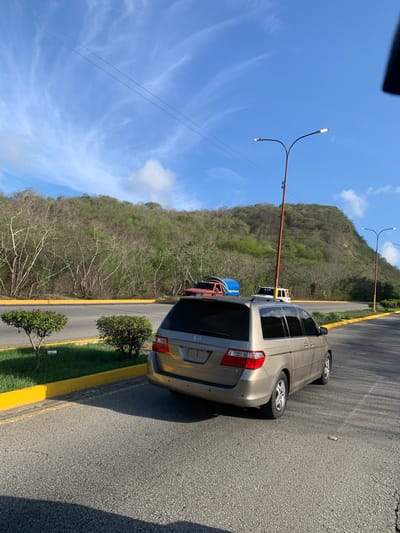 A tan Honda Odyssey minivan is parked on an asphalt surface, likely a parking lot, next to a grassy median with two small, manicured bushes. A yellow curb separates the grass from the asphalt. In the background, a lush, green, and wooded hillside rises under a bright blue sky with sparse, wispy white clouds. Two streetlights with orange poles are visible. On top of a red truck, which is partially obscured by the minivan, there is a blue object and a white SUV. The scene appears to be outdoors, possibly in a suburban or semi-rural area, given the presence of the natural landscape and the parking lot infrastructure. The photograph is taken from a slightly elevated perspective, looking towards the minivan and the hill.