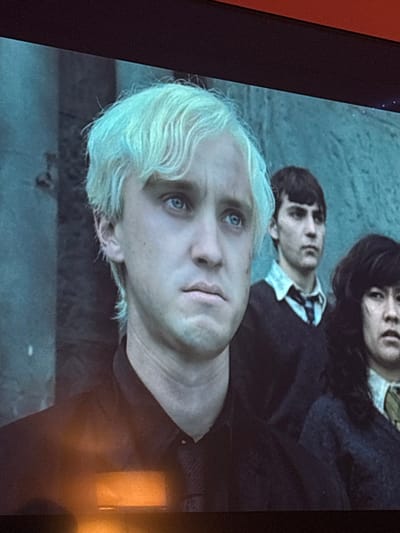 The image shows Draco Malfoy, with platinum blond hair and pale skin, lying down and looking up with a concerned expression. Behind him are two other figures: one with dark hair and a dark school uniform, and another with short brown hair and a white shirt with a tie. The background is blurred, suggesting they are lying on a surface. The overall tone is serious and somewhat somber.