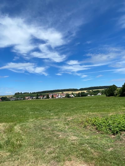 An outdoor, eye-level, vertical, medium shot captures a sunlit rural landscape under a bright blue sky with scattered white clouds. In the foreground, a vast, green field with patches of dry, brown grass dominates the scene. A faint path is visible on the left side of the field. In the middle ground, a village with red-roofed houses and white facades is nestled amongst trees and rolling hills. The village appears to be Christerode, Germany. The background features a dense line of dark green trees atop the hills, with a lighter green and yellow field visible behind the village. The overall impression is one of a serene, pastoral environment.