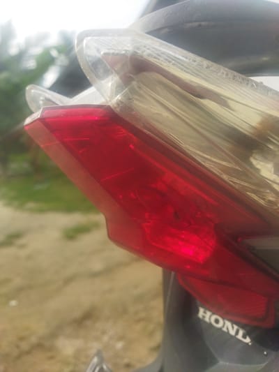 The media file shows a close-up, angled shot of a red motorcycle tail light. The tail light is clear red plastic with internal red reflectors and bulbs visible. Above the red section is a clear, ribbed plastic lens, likely for the turn signals or brake light. A transparent plastic film is still wrapped around part of the tail light assembly, suggesting it might be new or recently acquired. Below the tail light, the black plastic body of the motorcycle is visible, with the white "HONDA" logo clearly imprinted on it.

The background is out of focus, showing a blurred expanse of light brown ground with patches of green vegetation, hinting at an outdoor setting. Tall, slender trees with green foliage are faintly visible in the upper left background. The lighting suggests it is daytime, likely with bright sunlight creating some glare on the plastic surfaces. The overall scene appears to be in a rural or semi-rural location in Lancokpanteara, Indonesia. No people or specific activities are depicted. The focus is entirely on the motorcycle's tail light and branding.