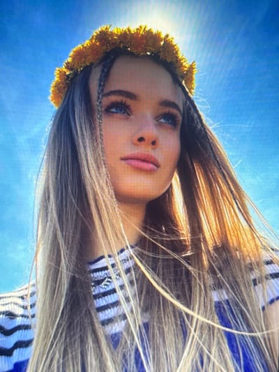 A low-angle medium shot shows a young woman wearing a crown of yellow flowers. She has long blonde hair, some of which is braided and falls over her shoulders. Her eyes are blue, and her lips are painted pink. She is looking upward and to the left, with the sun glinting behind her head. She is wearing a striped shirt with blue and white stripes and some shimmering embellishments. The background is a bright blue sky. The image appears to be taken outdoors, possibly in a park or field, in Shcherbanka, Ukraine.