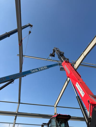 The image captures an outdoor construction scene under a clear blue sky in Stróża, Poland. The primary focus is on the assembly of a steel structure, with large gray I-beams forming the framework of a building. Two cranes are actively engaged in lifting and positioning these beams.
On the left, a black crane arm extends upwards, holding a steel beam suspended by red and green straps. A yellow and black hook mechanism is visible at the end of the crane's cable. Adjacent to this crane, a blue boom lift is positioned, displaying the text "NER MASZYNY Genie Z-80/60".
To the right, a large red telescopic crane, identified by the "MANITOU" logo and "WELDdetail TH group" branding, is prominently featured. Its extended boom is angled upwards, reaching towards the steel beams. A portion of a red construction vehicle, likely the base of the Manitou crane, is visible at the bottom of the frame, showing a black cab with a yellow flashing light. The steel beams are connected at various points, indicating ongoing construction progress. The ground is not visible in the image, but the overall impression is one of active construction work in an open area.