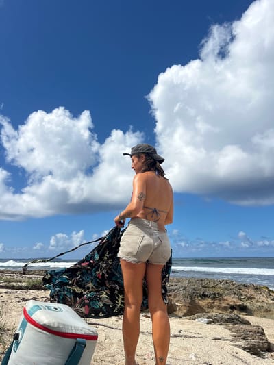 An adult female stands on a sandy and rocky beach in Le Moule, France, depicted from behind and slightly to her right, looking towards the ocean. She wears a black baseball cap, a patterned bikini top with visible ties at the back, and light tan denim shorts. Tattoos are visible on her left bicep and lower left leg. She holds a large, dark-colored fabric garment with teal leaf and pink floral patterns, which appears to be unfurling or moving in the wind. A white and red soft-sided cooler bag with blue accents and a snowflake logo rests on the sand in the lower left foreground. The environment features a sandy foreground with sparse dry vegetation, transitioning to a rugged rocky shoreline that meets a blue ocean with breaking waves. The sky is bright blue, marked by large, scattered white cumulus clouds.