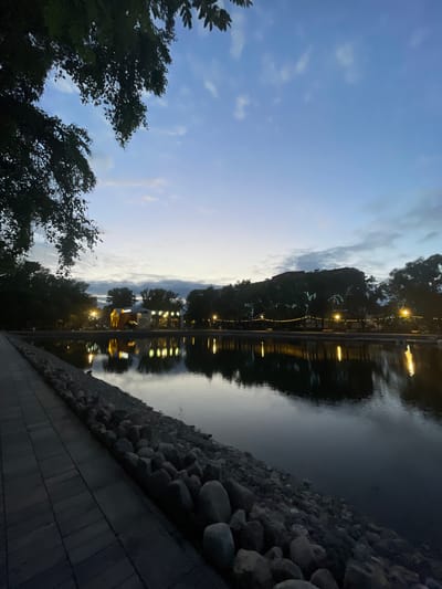 This photograph captures a twilight scene in Vologda, Russia, at the city's central pond. The viewpoint is from a paved walkway that runs along the pond's edge, lined with large stones. On the opposite bank, trees are silhouetted against the darkening sky, with lights from buildings and string lights illuminating the foliage. The pond reflects the lights and the sky, creating a shimmering effect on its surface. The sky is a gradient of deep blue and lighter shades, with wispy clouds scattered across it. The overall atmosphere is calm and serene, suggesting the end of a day.