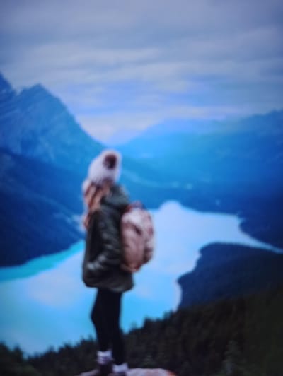 The image is a blurry, vertical shot of a person from the back, standing on a rocky outcrop overlooking a vast, mountainous landscape with a vibrant turquoise lake. The person is wearing a winter hat with a pom-pom, a dark puffer jacket, black leggings, and white socks with ankle boots. They are carrying a backpack. The background features imposing mountains, a body of water that appears to be a lake, and a cloudy sky. The scene depicts a moment of solitary contemplation in nature. The location is specified as Balakliya, Ukraine.
