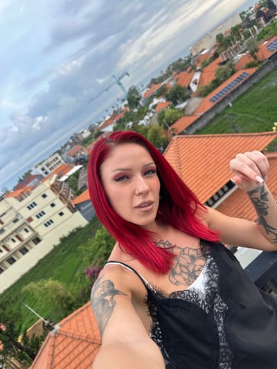 A woman with red hair is photographed outdoors with a cityscape visible in the background. She is wearing a black top with lace detail over a white garment, and her arms and chest are tattooed. She is holding up her thumb. In the background are buildings with orange roofs, greenery, and a construction crane. The location is Tibubeneng, Indonesia.