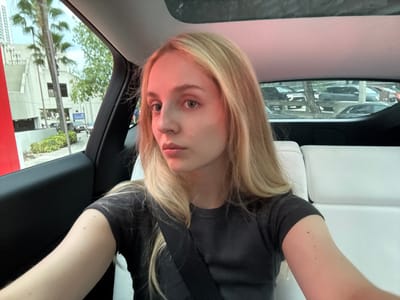A young Caucasian female with blonde hair and blue eyes is seated in the back seat of a car, looking at the camera. She is wearing a dark gray t-shirt and a black seatbelt. The car's interior is visible, with white leather seats and a sunroof. Outside the car, a cityscape with buildings and palm trees can be seen through the window, suggesting a location in Miami, Florida, United States of America. The lighting suggests it is daytime.