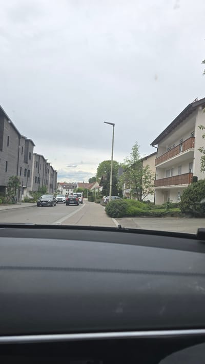 The photograph shows a view from inside a vehicle driving on a street in what appears to be a residential area. The buildings on the right side of the street appear to be apartment buildings with balconies, while the buildings on the left side are modern, grey apartment buildings. Several cars are visible on the road ahead. A streetlight is seen on the left side of the street. The weather appears overcast. The location is Pfaffenhofen an der Ilm, Germany.