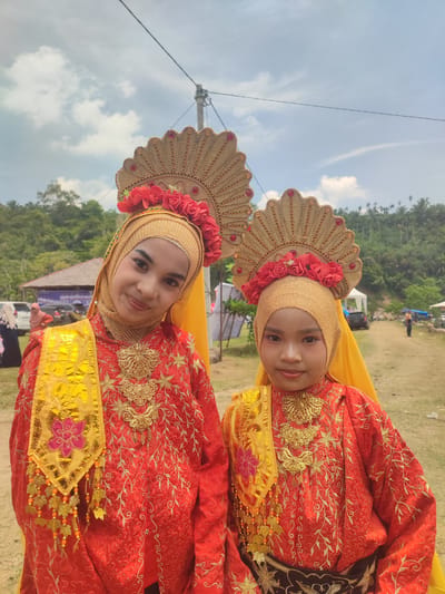 Two young girls, one appearing older than the other, are posing outdoors in traditional Indonesian attire. Both are wearing gold hijabs and elaborate headdresses adorned with red flowers. Their red dresses are heavily embroidered with gold thread and feature gold necklaces and decorative sashes. The older girl is on the left, looking towards the viewer with a slight smile, while the younger girl is on the right, also facing the camera. In the background, there are trees and rolling hills, suggesting a rural or semi-rural setting. Several tents and vehicles are visible in the distance, and people can be seen milling about, indicating an event or gathering. The sky is partly cloudy. This scene is located in Pantonrayeuk Satu, Indonesia.