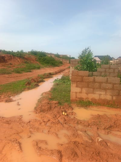 The media file depicts a muddy construction site in Reifild-Ist, Nigeria. The scene is dominated by reddish-brown mud and puddles of murky water, suggesting recent rainfall. A dirt path winds through the site, partially submerged in water. On the right side of the frame, a partially constructed wall made of concrete blocks stands, with some overgrown vegetation emerging from cracks. In the background, more unfinished structures and scattered trees are visible, along with glimpses of houses with tiled roofs, indicating a nearby residential area. The sky is a pale, hazy blue, hinting at an overcast or early morning/late afternoon time of day. The overall impression is one of ongoing construction work in a natural environment that is currently experiencing wet conditions. There is no visible text in the media file.