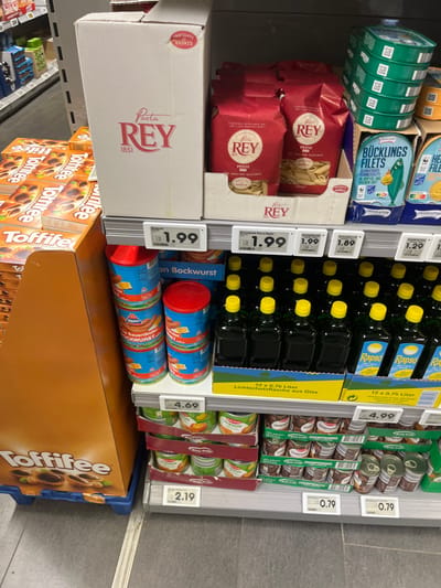 The image shows a grocery store shelf with a variety of food items. The shelf is located in a supermarket at the coordinates 50.919, 6.923, which corresponds to Cologne, Germany.  The shelf is organized by product category with the top shelf displaying cans of fish labeled "Bücking's Filets".  The next shelf down displays boxes of pasta labeled "Rey".  The third shelf contains bottles of oil with yellow lids and "Rapso" printed on them.  Below the oil bottles are two rows of red-lidded metal cans of "Bockwurst".  The bottom shelf is a display of "Toffee" candy.  All the products are on sale and have price tags attached.