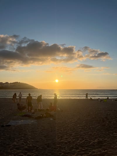 This photograph depicts a sunset scene on a beach in A Coruña, Spain. The sun, a bright orb, is low on the horizon, casting a golden glow across the sky and sea. Several people are scattered across the sandy beach, some standing, others sitting or lying down on towels. They appear to be enjoying the evening. In the background, the city of A Coruña is visible as a silhouette against the brightly lit sky. The presence of the urban skyline, coupled with the specific beach setting and the characteristic light of a sunset in this region, strongly suggests the location is likely Playa de Riazor or Playa del Orzán, prominent urban beaches within A Coruña. The image captures a moment of relaxation and natural beauty.