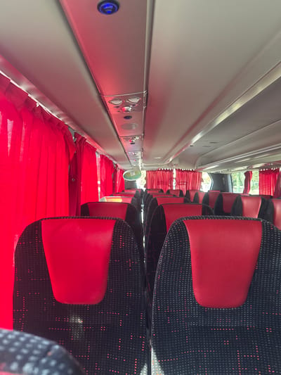 The image shows the interior of a bus, likely in Bnei Brak, Israel. Rows of red and black patterned seats are visible. Above the seats are individual overhead consoles with buttons and lights, likely for passenger use. Red curtains line the windows. The perspective is angled, giving a partial view of the bus's interior.