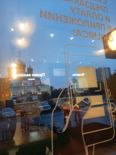 The image shows a reflection on a glass surface, likely a shop window. The reflection reveals a street scene in Barnaul, Russia, at dusk. In the background, a prominent Russian Orthodox church with golden domes is visible, along with modern buildings. The foreground of the reflection includes images of cars, a street, and people. There are also graphical elements on the glass, possibly advertisements or branding. Text, reversed due to reflection, can be seen on the glass, with Cyrillic characters. Specifically, text appears to read "ПРЕДЛОЖЕНИЯ," "УПАКОВКА," and "В НАЛИЧИИ," indicating offers, packaging, and availability. Another prominent reflection shows a graphic of a smartphone and a hand holding it, suggesting a technology-related business. The street appears wet, indicating recent rain.