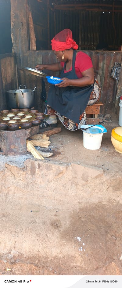 In the open-air setting of Barikin Gidar Kanat, Nigeria, a woman wearing a red headscarf and a red shirt, with a black apron over her patterned skirt, is actively preparing food. She is seated on a low wooden stool, facing a traditional cooking setup. In front of her, a round, cast-iron cooking mold with many small, cup-like depressions is filled with what appear to be small, cooked cakes or pancakes, situated over burning embers. To her left, a large, silver pot with two handles rests on a metal structure, also over the heat source. The woman holds a blue plastic dish in her left hand, containing food items, while her right hand is positioned over the cooking mold as if tending to the food or adding to it. The environment is rustic, with a dirt floor and what appears to be corrugated metal sheeting forming a wall or roof in the background. Various objects are scattered around, including pieces of firewood, a white plastic bucket with a blue lid, and another yellow container. The overall scene depicts a domestic food preparation activity in a rural Nigerian context.
