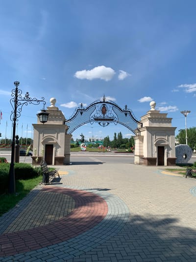 This is a low-angle, vertically oriented, full shot of an ornate archway entrance to a park or public space on a sunny day. The sky is a vibrant blue with scattered white clouds. The arch is made of light-colored stone and features intricate black metalwork. The metalwork includes a decorative crest and what appears to be text. Below the arch, a paved walkway of light gray, pink, and blue bricks leads into the park. Green grass and trees line the walkway, and there are several decorative lampposts with black ironwork. A bench is visible in the foreground, casting a shadow on the paving stones. In the background, across a road, more trees and buildings are visible, along with a statue of a person on horseback. The overall scene suggests a pleasant day in a well-maintained urban park. The text on the arch reads "ТИРАСПОЛЬ", indicating the location is Tiraspol. The time of day appears to be late morning or early afternoon, given the bright sunlight and the shadows cast. There are no people prominently featured in the image.