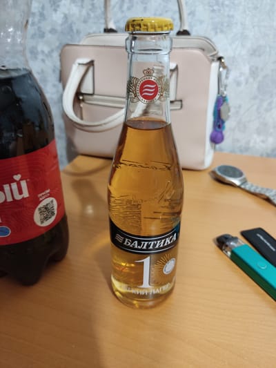 A close-up, eye-level shot captures a wooden surface with various objects. In the foreground, a clear glass bottle of golden liquid, labeled "БАЛТИКА 1", stands prominently. To the left, a dark plastic bottle with a red label featuring a white heart icon is partially visible. Further back, a light-colored handbag with white handles and a keyring adorned with colorful beads rests against a textured wall. The scene includes a metallic spoon and a teal electronic cigarette device with a black accessory. The lighting appears to be artificial, casting soft shadows. There is no discernible information to determine the location as Serpukhov, Russia, as the image lacks any identifying landmarks or contextual clues specific to that city.