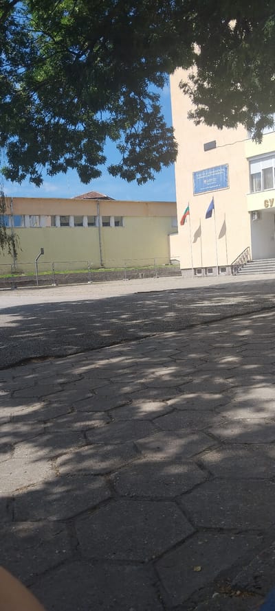 This is an exterior, eye-level, medium-range shot of a school building and its grounds in Svishtov, Bulgaria. The building is light yellow with a tan roofline. There is a sign on the front of the building that says "CAMBRIDGE SCHOOL" in English and in Bulgarian. There are three flags flying in front of the building: a Bulgarian flag, and two other flags that are dark blue. The grounds in front of the building are paved with hexagonal concrete pavers, and there are shadows from trees cast on the ground. To the left, there is a low, yellow building with a tan roof and several windows. A chain-link fence runs in front of this building. The sky is blue with some clouds.