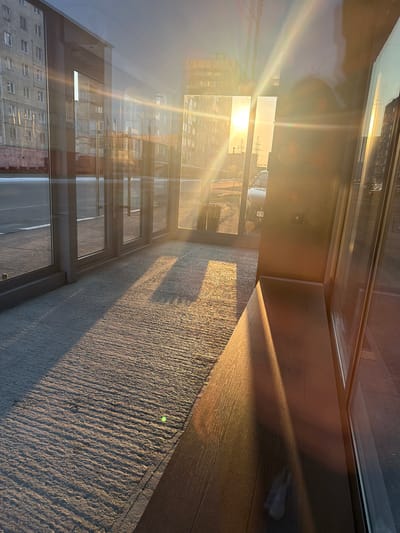 This image captures a sun-drenched interior space, likely a room or hallway with large windows. The sun is low in the sky, casting long, dramatic shadows across the textured floor and wooden surface. The strong sunlight creates lens flares and bright highlights, suggesting a late afternoon or early morning sun. Through the windows, an urban landscape is visible, featuring multi-story buildings with visible window panes. There are also signs of industrial infrastructure, with what appear to be power lines or transmission towers in the distance, consistent with an industrial setting. The reflections in the glass also show parts of buildings, adding to the urban context. The overall mood is tranquil and serene, bathed in the warm glow of the setting or rising sun. There is no visible text in the image.
