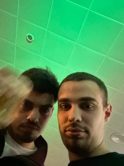 Two men appear to be in a room with a tiled ceiling. The ceiling is lit with a green hue. A circular vent is visible on the ceiling. The location is not specified.