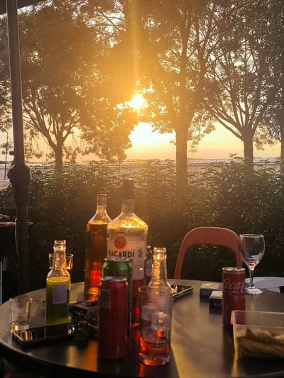 A round table outdoors, set with various bottles of alcohol and drinks, is bathed in the warm, golden light of a sunset. The scene appears to be on a patio or garden area, with lush green bushes in the background and large trees silhouetted against the bright sky. The sun is prominently visible, peeking through the branches of the trees, indicating it is late afternoon or early evening. The overall mood is relaxed and convivial, suggesting a casual gathering.
On the table, there are several bottles, including a bottle of Bacardi rum, another with amber-colored liquid, and a clear bottle with green liquid. Cans of Coca-Cola and another beverage are also present. Empty and partially filled glasses, including a wine glass, are scattered on the table, along with a mobile phone, a pack of cigarettes, and a small container of food. The reflection of the setting sun creates a beautiful glow on the table surface and the objects. The presence of alcohol and drinks suggests a social occasion, possibly a get-together with friends or family. The location context of Almere, Netherlands, places the scene in a Dutch setting, likely a residential area or a casual dining establishment. The weather appears to be clear, allowing for an unobstructed view of the sunset.