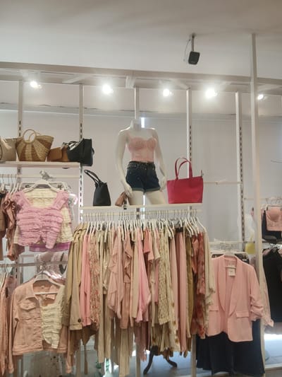 The image depicts a clothing store display. In the foreground, clothing items hang on racks, predominantly in shades of pink and beige. There are several garments including jackets, tops, and dresses. Some of the tops have ruffled details, and one jacket is a light pink denim material. In the middle ground, a mannequin is displayed wearing a pink corset top and dark shorts with sequin embellishments. To the left of the mannequin, there are several handbags on shelves, including a straw bag and a black leather bag. A bright red tote bag is also visible on a shelf. The background shows more shelves and clothing racks. The overall environment appears to be a retail store with bright overhead lighting and white shelving units.
