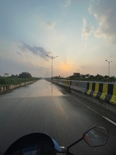 The image captures a serene, post-rain scene along a highway in Purbachal, Bangladesh, likely during sunset or sunrise. The perspective is from a vehicle, with a portion of its handlebar and rearview mirror visible in the foreground on the right.
The central feature is a wide, wet road stretching into the distance, reflecting the soft light of the sky. A prominent median barrier separates the road, painted in alternating black and yellow stripes. To the left of the barrier, a strip of green foliage and distant trees line the road. Numerous streetlights with distinct double-arm designs are spaced along the median.
The sun, a bright circle with visible rays, is low on the horizon, positioned centrally-left within the frame, casting a warm glow across the sky. The sky itself is mostly clear with scattered, light clouds, showing a transition from a pale blue to a warmer, golden hue near the sun. The wet road surface indicates recent rainfall, adding a reflective quality to the scene.
Notable details include:
* On the yellow and black median barrier, the number "100" is visible on a yellow section, and an emblem resembling crossed axes or hammers within a circle is seen further down.
* On the vehicle's dashboard or display, the number "26" is clearly visible.
* The rearview mirror on the right reflects a blurry image of the surroundings, including possibly a person's arm.
* What appear to be distant wind turbines or tall poles are visible near the horizon to the right of the sun.
The overall atmosphere is tranquil and highlights the natural beauty of the sky and the infrastructure of the road, suggesting a peaceful drive after a rain shower.