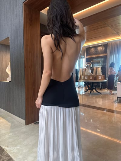 The image shows a female individual, viewed from the back, positioned in an indoor commercial or event space located in Monterrey, Mexico. The subject has long, dark, wavy hair and is wearing a black, backless top or dress featuring a visible zipper running vertically down the spine, paired with a long, white, pleated skirt. Her right arm is slightly raised, with her hand positioned near the back of her head.
To the left, a grey textured wall panel is visible alongside a warm-toned wooden door frame. Beyond this frame, a white, abstract sculptural object rests on a dark surface. The floor throughout the visible area consists of highly polished light-colored marble or stone tiles, reflecting ambient light.
The background reveals a spacious, well-lit interior with a wooden panel ceiling equipped with linear recessed lighting. Large windows with sheer curtains line the right side, allowing natural light to enter. A dark, possibly metallic or mirrored, wall feature is present. In the mid-ground, a round table with a decorative dark base displays several white-wrapped items and a reflective metallic object resembling a helmet. Two other females are partially visible in the background; one in a light-colored patterned top appears to be observing items near the table. Additional clothing racks are discernible further in the background.