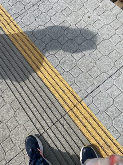 The image shows a ground-level perspective of a textured pavement area, likely a sidewalk or pathway, in Lahošť, Czechia. The pavement is composed of light gray, interlocking hexagonal tiles with a rough surface. A raised, bright yellow tactile paving strip runs diagonally across the frame, separating two distinct sections of the pavement. To the left of the yellow strip, the pavement tiles are oriented with a pattern of parallel raised lines. To the right of the yellow strip, the hexagonal tiles are laid in a more traditional pattern, with a large, irregular shadow cast across them from an unseen object.
In the lower portion of the image, two individuals are visible from the knees down, wearing footwear. On the left, a person is wearing dark blue athletic shoes with white soles and pink accents. On the right, another person is wearing dark blue athletic shoes with white soles. The individuals appear to be standing still. The overall scene suggests an outdoor urban or public space, possibly near a street or building, given the paving and the presence of a tactile warning surface. The lighting indicates a sunny day, with distinct shadows.