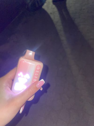 A close-up, over-the-shoulder shot captures a hand with manicured nails holding a pink vape device. The device has a light pink and white gradient design with glowing cartoonish figures and the text "ELFBAR" visible. A soft blue light emanates from the top of the vape. The background is a dark, out-of-focus surface, possibly asphalt or concrete, with subtle shadows suggesting an outdoor, urban setting. The overall lighting is dim, with a strong highlight on the vape device. This image was taken in Velykyi Buialyk, Ukraine.