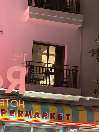 This media file shows a close-up view of a building facade in Sant Antoni De Portmany, Spain. The building is illuminated with pink and yellow lights, casting a warm glow on the scene. On the left side of the image, a large pink sign is visible, with the word "THE" in white letters and a large pink letter "R" below it. To the right of the sign, a colorful vertical sign displays the word "SUPERMARKET" in red letters against a background of alternating yellow and blue stripes. A bright red neon light runs along the right edge of the "SUPERMARKET" sign. Above the "SUPERMARKET" sign, a small white box with text is partially visible, and the words "Hielo ICE CUBES" are legible.

The building itself has a modern design, with white architectural elements and a balcony area with dark railings. A window on the balcony shows interior lighting and hints of furniture. The upper portion of the image is partially obscured by the leaves of a tree. The overall impression is of a commercial building, likely a hotel or a establishment with a supermarket and possibly a hotel component. The time of day appears to be evening or night due to the artificial lighting. The scene is brightly lit and likely bustling with activity, given the prominent signage.