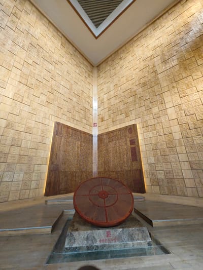 A ground-level, indoor shot features a large, circular object displayed on a pedestal within a building in Daxing District, China. The building's walls are covered in square tiles, each bearing inscribed Chinese characters. Behind the pedestal is a large, open book-like structure also inscribed with Chinese text, illuminated by lights at its edges. The floor is made of large, light-colored tiles, with steps leading up to the display area. The ceiling has a rectangular opening with a view to a higher level featuring a slatted design.