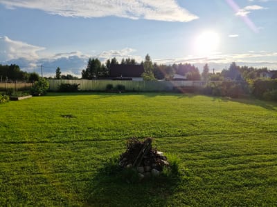 This image depicts a sunlit lawn in the village of Sorochkino, Russia. The foreground features a neatly mown green lawn with a small pile of kindling and stones in the center. A metal fence runs across the middle ground, separating the lawn from a wooded area and a few residential buildings. The sky is blue with scattered white clouds, and the sun is bright, casting long shadows across the grass. The overall scene is tranquil and rural, suggestive of a summer evening.