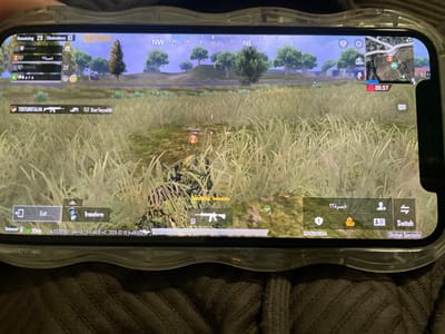 The image displays a mobile phone screen showing the PUBG Mobile game. The player is in a grassy field, seemingly hiding. The HUD displays remaining players, team information, a mini-map with a timer, and various in-game controls like "Exit," "Transform," and weapon switch options. Two usernames: "TSKTURKTALHA" and "Star Tayyabi" are visible, possibly representing teammates. The phone is in a protective case.