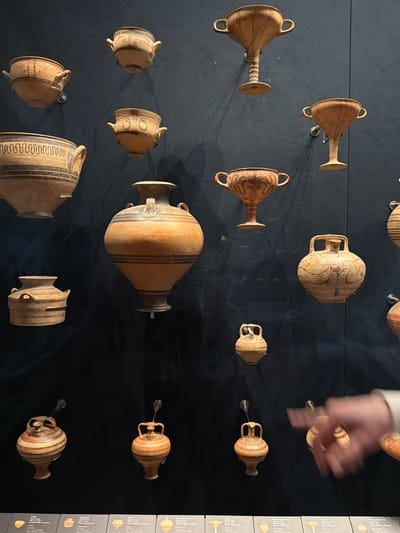 A museum display features a collection of ancient Greek pottery mounted on a dark blue wall. The pottery varies in size and shape, including amphorae, kraters, and kylixes, all exhibiting intricate decorative patterns in earth tones. The items are evenly spaced and appear to be illuminated by spotlights, creating subtle shadows. In the lower right portion of the image, a person's hand is partially visible, blurred by motion as it reaches towards the display. Below the pottery, a label strip with text is visible, suggesting this exhibit is located in Istanbul, Turkey.