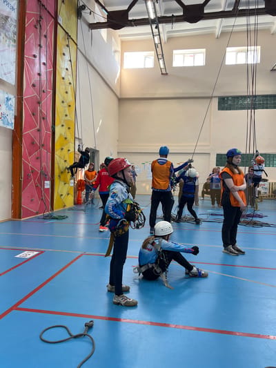 Several people are participating in an indoor rock climbing activity at a climbing wall in Berezniki, Russia. The climbing wall is divided into sections of different colors including red and yellow. Participants are wearing climbing harnesses and helmets, with ropes attached for safety. Some are actively climbing, while others are standing on the floor, likely waiting their turn or assisting. The indoor space has a blue floor marked with red lines. Large windows are visible in the upper part of the wall. Banners hang near the wall’s base.