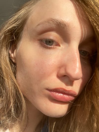 Close-up portrait of a fair-skinned person with light blonde hair, tilted to the side. The person has gray-green eyes, a small nose, and full lips. The skin appears smooth and has a slight sheen, possibly due to lighting or moisturization. The overall scene appears to be indoors with natural lighting, casting shadows across the face. There is no information available from the image or the context provided that indicates the location is Sepych, Russia.