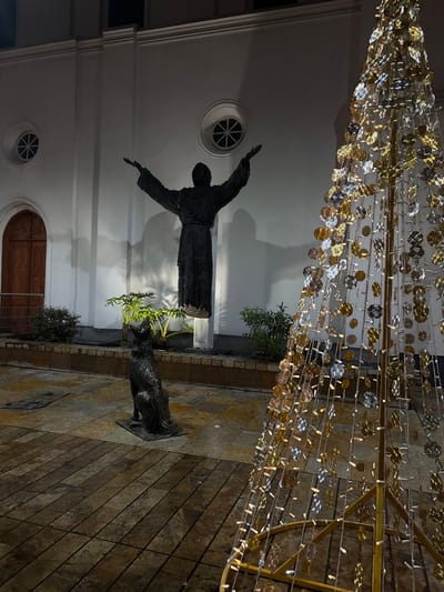 A bronze statue of a robed figure with outstretched arms stands before a white building with two round windows and an arched doorway. A bronze dog statue sits near the statue. A decorative structure with shimmering discs and lights hangs above. The scene is lit by artificial lighting, casting shadows and highlighting the textures of the statues and building. The ground is paved with stone tiles.