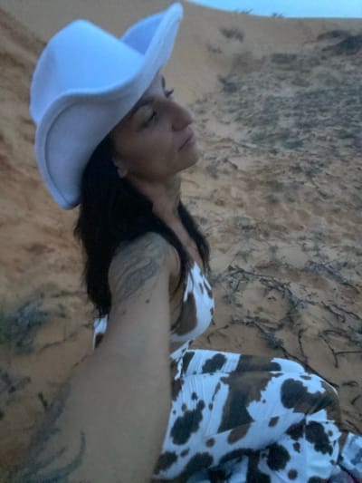 A woman wearing a white cowboy hat and a cow-print outfit is sitting on sand dunes. Her eyes are closed and her head is tilted back as if she is enjoying the sun. She has tattoos on her arm. The sand dunes are a light orange color and there are some dry plants scattered around. The sky is a light blue. The location is the sand dunes of Mui Ne, Vietnam.