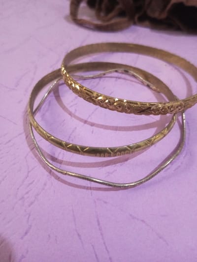 The scene is located in the city of Chandragiri, Nepal. The image displays a close-up, slightly elevated view of three circular bangles resting on a light purple, subtly textured surface. The uppermost bangle is gold-toned, approximately 5mm wide, and features an intricate, embossed pattern composed of alternating stars and rounded geometric designs. Positioned directly below it is a thinner, gold-toned bangle, approximately 3mm wide, adorned with a repeating pattern of small diamond shapes and dots. The lowest bangle is silver-toned, the narrowest at roughly 2mm wide, and exhibits a slightly irregular, wavy circumference with minor surface wear. In the upper right background, a blurred, dark brown fabric object is partially visible. No individuals are present, and no actions or interactions are depicted.