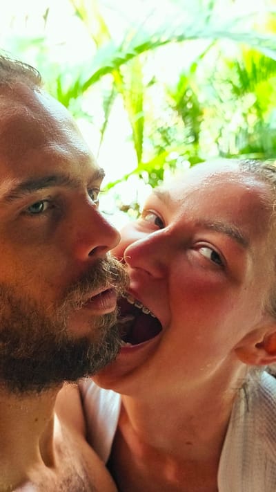 The image presents a tight, vertical close-up of two individuals in a playful, intimate interaction against a brightly lit, blurred background of green foliage. On the left, a male subject is partially visible from the forehead to the mid-chest, featuring a light brown beard, a prominent nose, and blue-green eyes looking slightly upward. His skin appears moist or sweaty. On the right, a female subject is visible from the forehead to the shoulder, with her mouth wide open, revealing orthodontic braces on her upper teeth and her tongue. Her eyes are wide open, directed towards the male, and her cheeks are flushed. Her skin also appears moist. The subjects' faces are in extremely close proximity, with the female's open mouth positioned near the male's beard/chin area. The background is an out-of-focus mass of vibrant green leaves, indicative of dense tropical or subtropical vegetation. The specific location within Mù Cang Chải, Vietnam, is not visually discernible from the close-up subjects and blurred background foliage.